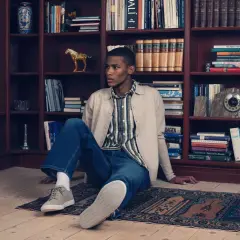 Ung man i avslappnad outfit sitter på en matta framför en bokhylla. En känsla av quiet luxury.