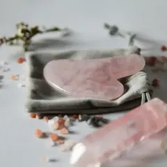 En rosa gua sha-sten ligger på en grå påse, omgiven av rosa saltkristaller och en kvartsstav.