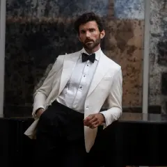 Man in a white tuxedo jacket and black pants leans on a black piano.