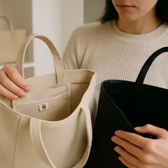 En person håller upp en ljusbeige och en svart väska. Den ljusa väskan har en liten logotyp med ett M. Perfekt för en vila väska.