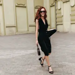 Woman with red hair in a dark green VILA midi dress, black sandals, and sunglasses, holding a small brown bag.