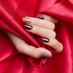 En hand med mörkröda naglar och en glittrig accentnagel vilar mot rött siden. En hyllning till red nail theory.