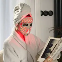 Person i morgonrock och handduk på huvudet läser en bok med en röd led mask ansikte.