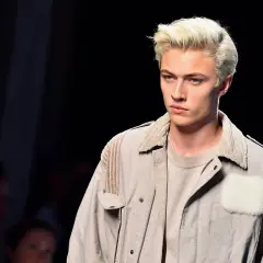 Lucky Blue Smith med blont hår och blå ögon på catwalken, klädd i en beige jacka med pälsdetaljer.