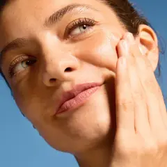Close-up of a woman applying serum to her cheek against a blue sky background.