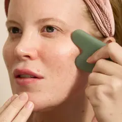 Kvinna med rosa pannband utför ansiktsyoga med en grön gua sha-sten för att massera kinden.