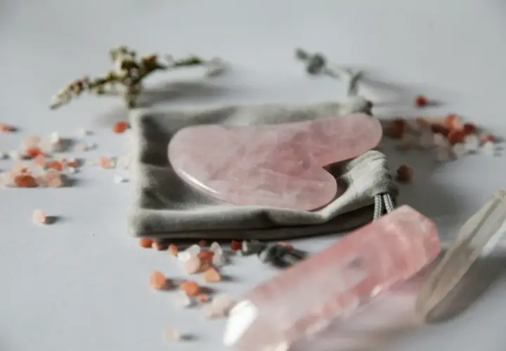 En rosa gua sha-sten ligger på en grå påse, omgiven av rosa saltkristaller och en kvartsstav.