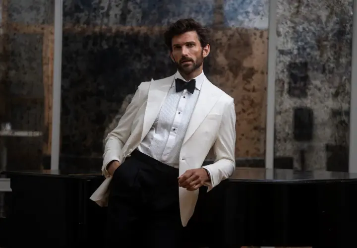 Man in a white tuxedo jacket and black pants leans on a black piano.
