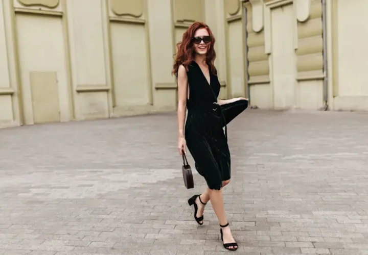Woman with red hair in a dark green VILA midi dress, black sandals, and sunglasses, holding a small brown bag.
