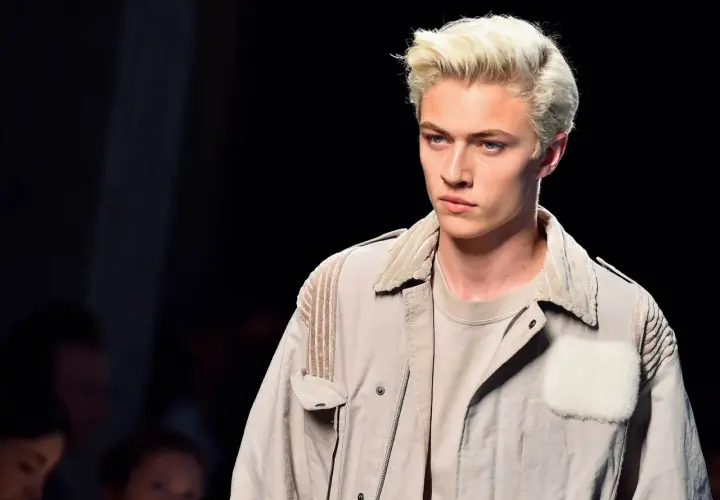 Lucky Blue Smith med blont hår och blå ögon på catwalken, klädd i en beige jacka med pälsdetaljer.