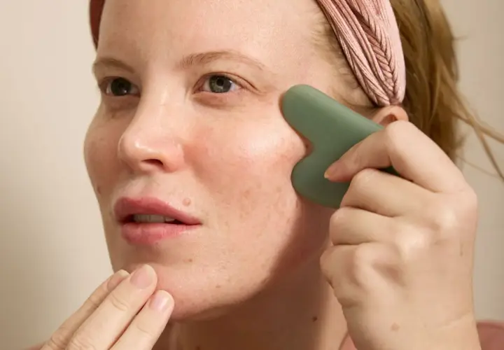 Kvinna med rosa pannband utför ansiktsyoga med en grön gua sha-sten för att massera kinden.