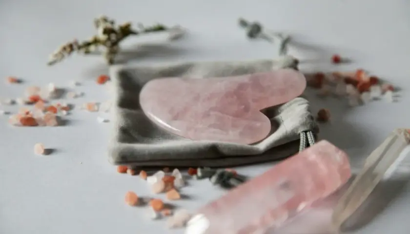 En rosa gua sha-sten ligger på en grå påse, omgiven av rosa saltkristaller och en kvartsstav.