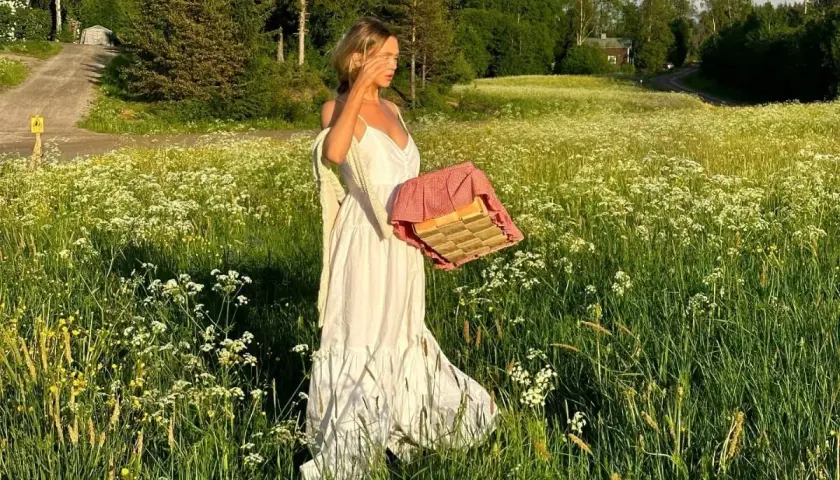 En kvinna i vita klänningar går genom en blomsteräng med en picknickkorg.