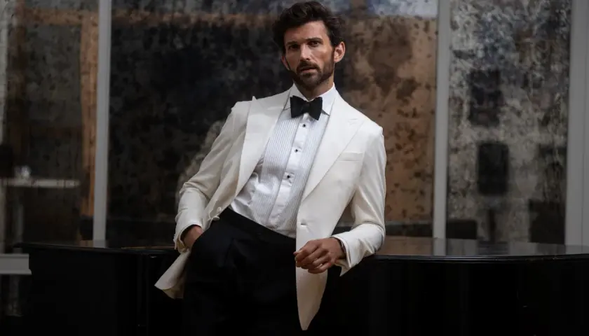 Man in a white tuxedo jacket and black pants leans on a black piano.