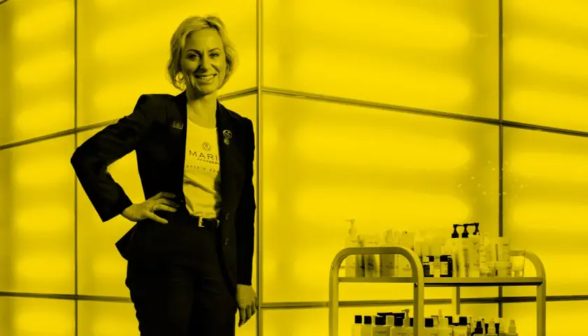 Maria Åkerberg, med en t-shirt med hennes namn, står stolt bredvid en hylla med hudvårdsprodukter.