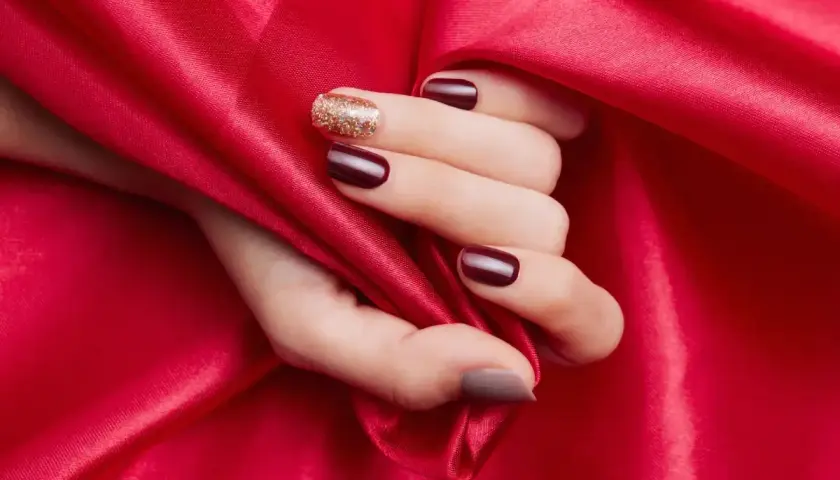 En hand med mörkröda naglar och en glittrig accentnagel vilar mot rött siden. En hyllning till red nail theory.