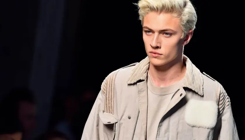 Lucky Blue Smith med blont hår och blå ögon på catwalken, klädd i en beige jacka med pälsdetaljer.