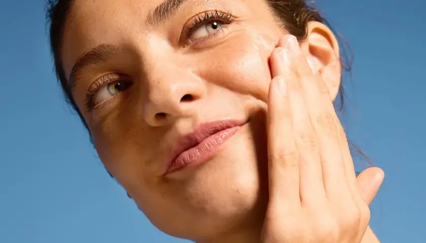 Close-up of a woman applying serum to her cheek against a blue sky background.