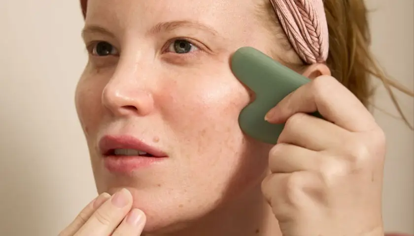Kvinna med rosa pannband utför ansiktsyoga med en grön gua sha-sten för att massera kinden.