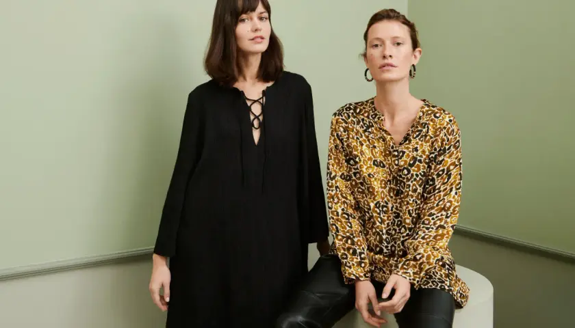 Två kvinnor poserar i en studio. Den ena bär en svart klänning med snörning, den andra en blus med leopardmönster. Båda utstrålar en elegant fransk stil.