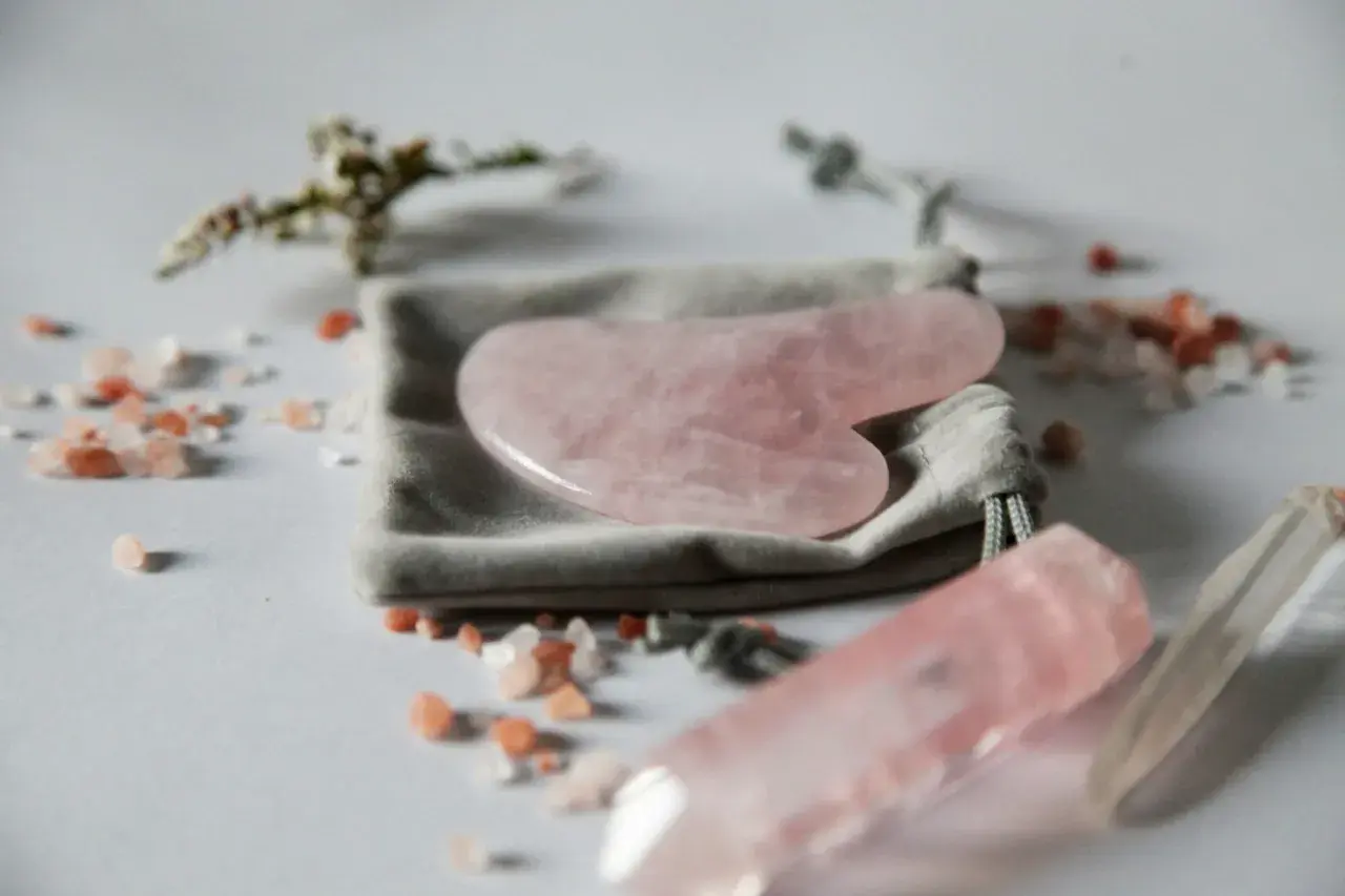 En rosa gua sha-sten ligger på en grå påse, omgiven av rosa saltkristaller och en kvartsstav.