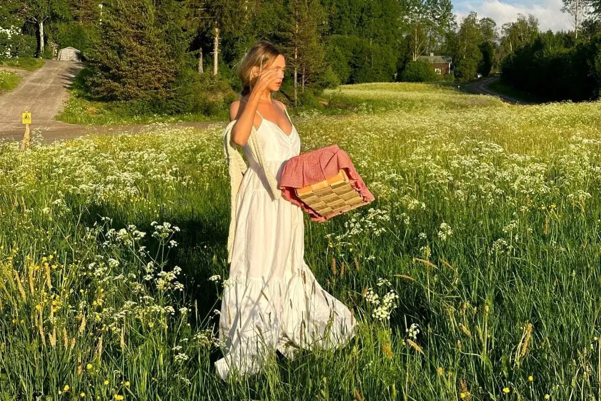 En kvinna i vita klänningar går genom en blomsteräng med en picknickkorg.