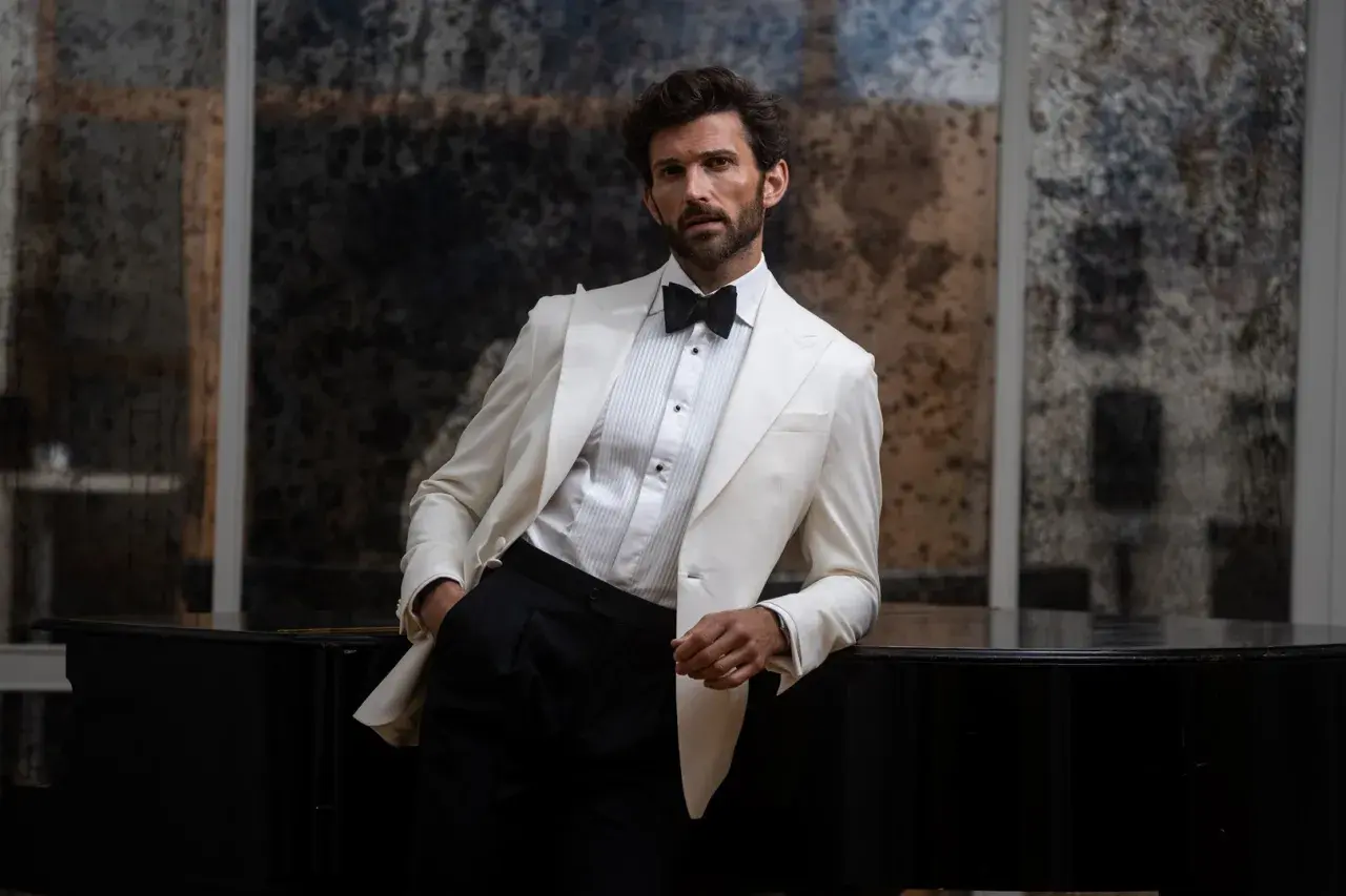 Man in a white tuxedo jacket and black pants leans on a black piano.