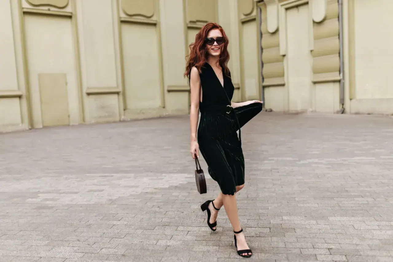 Woman with red hair in a dark green VILA midi dress, black sandals, and sunglasses, holding a small brown bag.