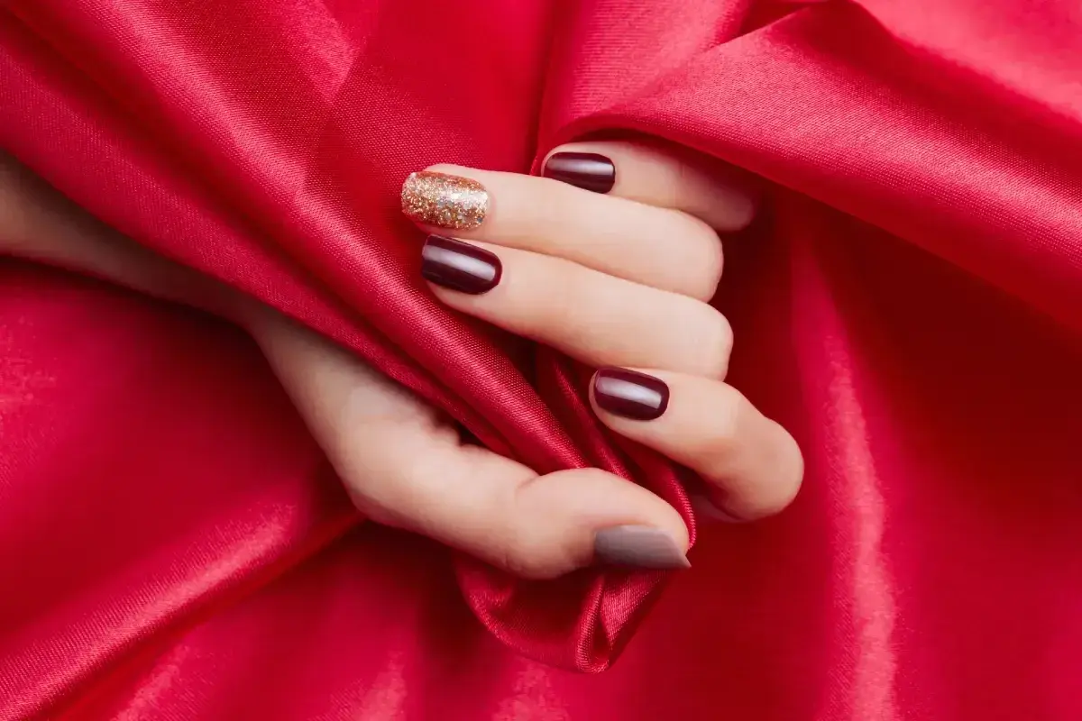 En hand med mörkröda naglar och en glittrig accentnagel vilar mot rött siden. En hyllning till red nail theory.