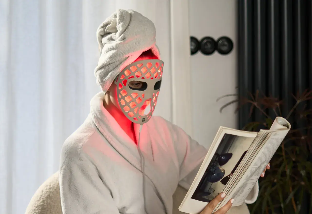 Person i morgonrock och handduk på huvudet läser en bok med en röd led mask ansikte.