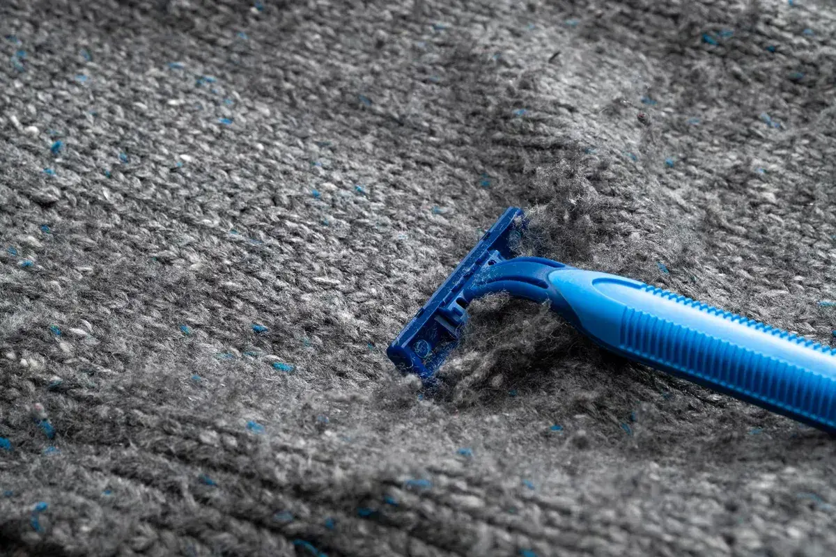 En blå rakhyvel ligger på en grå stickad tröja, redo att ta bort noppor.