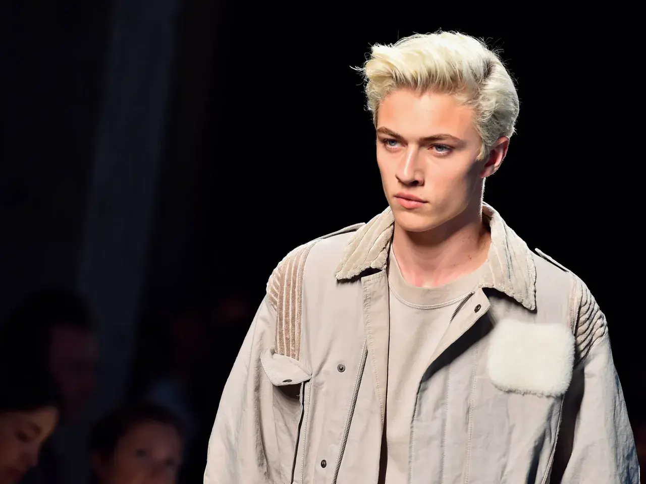 Lucky Blue Smith med blont hår och blå ögon på catwalken, klädd i en beige jacka med pälsdetaljer.