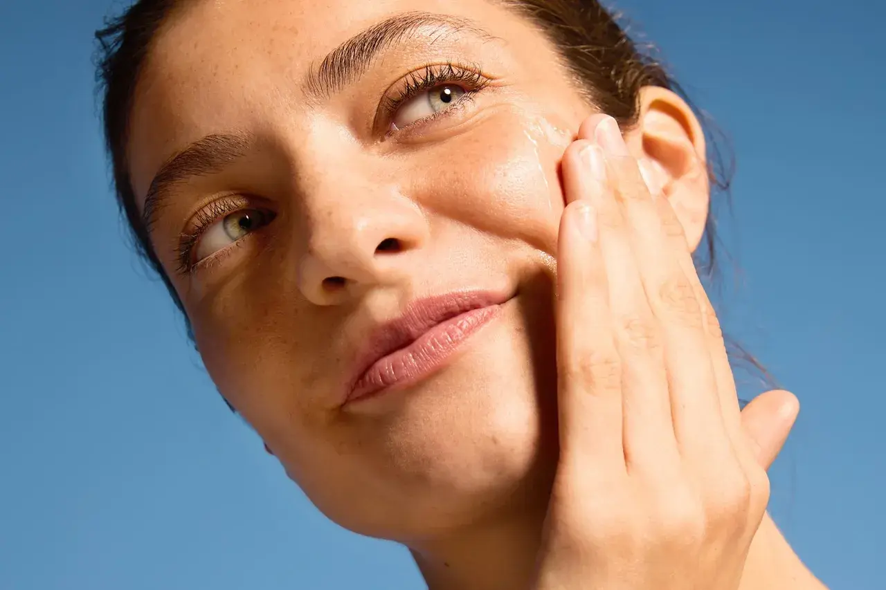 Close-up of a woman applying serum to her cheek against a blue sky background.