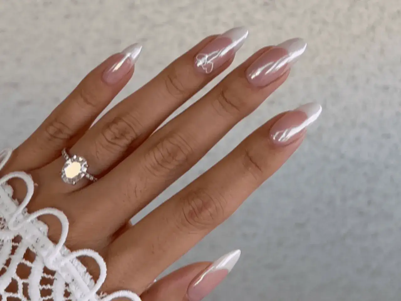 En hand med en glittrande ring och en elegant fransk manikyr med pärlemorskimrande toppar. En liten hjärtdekoration på en nagel.
