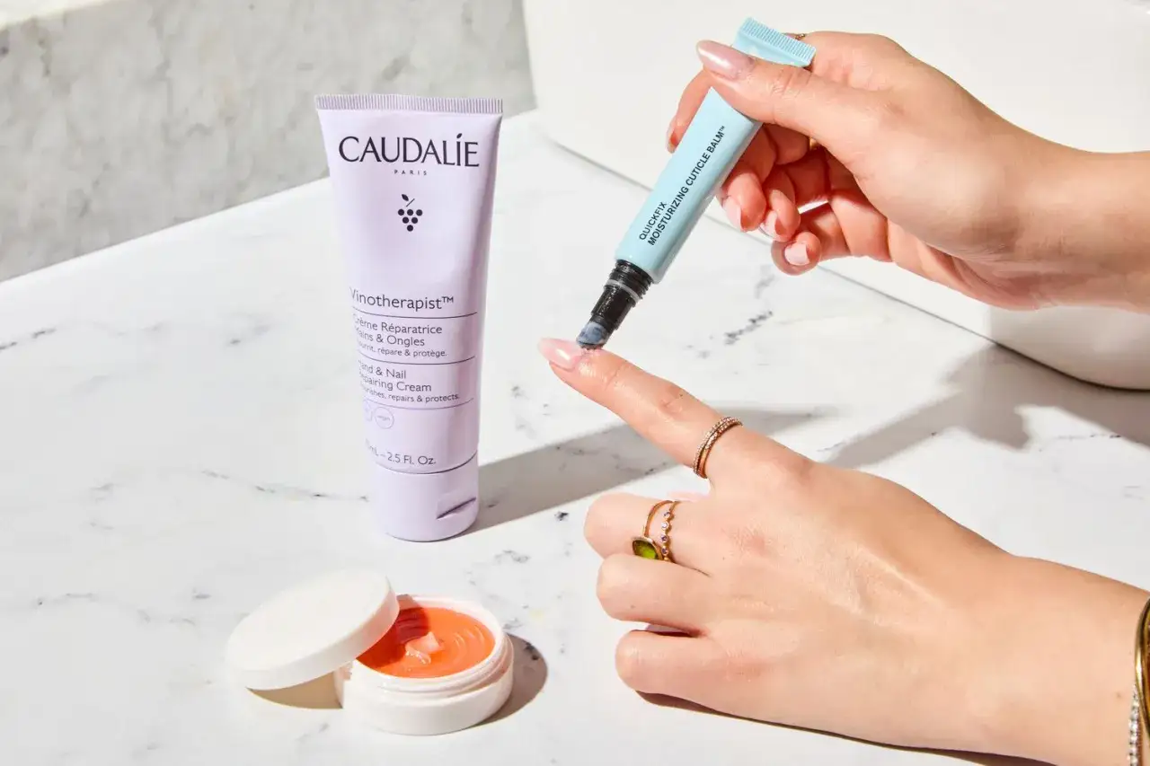 En hand applicerar nagelbandsolja för att behandla torra nagelband. En tub Caudalie handkräm och en burk med orange salva syns också.