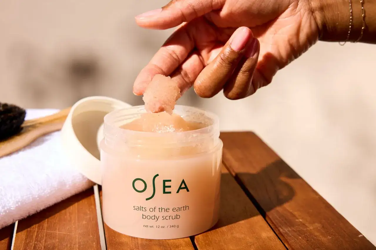 En hand tar en nypa av en rosa kroppsskrubb, en h&auml;rlig mekanisk peeling f&ouml;r huden. En vit handduk och borste syns i bakgrunden.