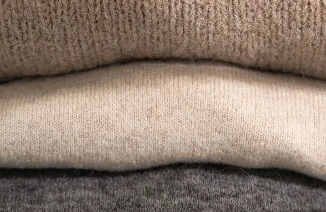 En hög med mjuka stickade plagg i olika nyanser av beige och grått. En av tröjorna har lite noppor som ger den en mysig känsla.