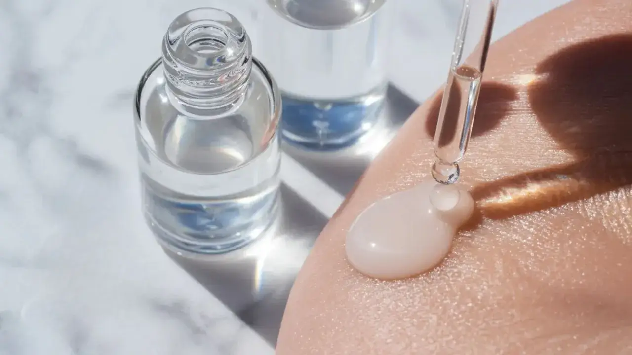 En droppe serum med sodium hyaluronate appliceras på huden från en pipett. Två flaskor med klar vätska står i bakgrunden.