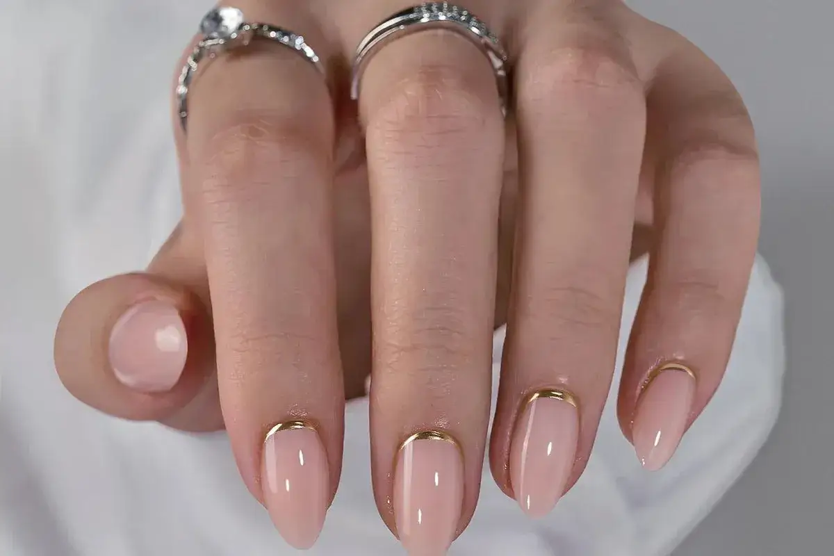 Enkla naglar med ljusrosa färg och en tunn gulddetalj vid nagelbandet. Vackra ringar pryder fingrarna.