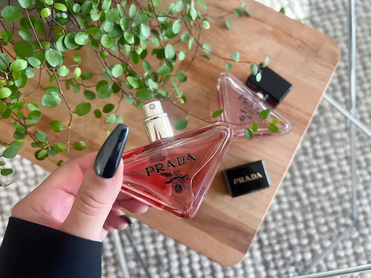 En hand med svarta naglar håller en Prada Paradoxe Intense parfymflaska. Doftnoterna av Prada Paradoxe Intense framträder i den rosa vätskan.
