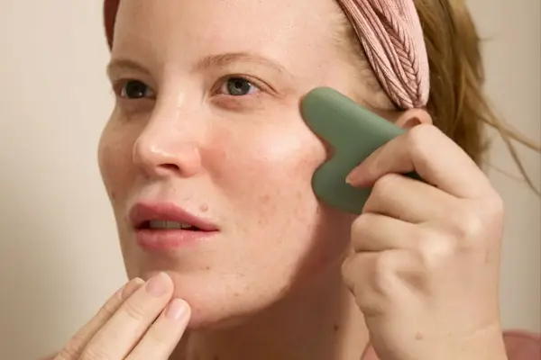 Kvinna med rosa pannband utför ansiktsyoga med en grön gua sha-sten för att massera kinden.